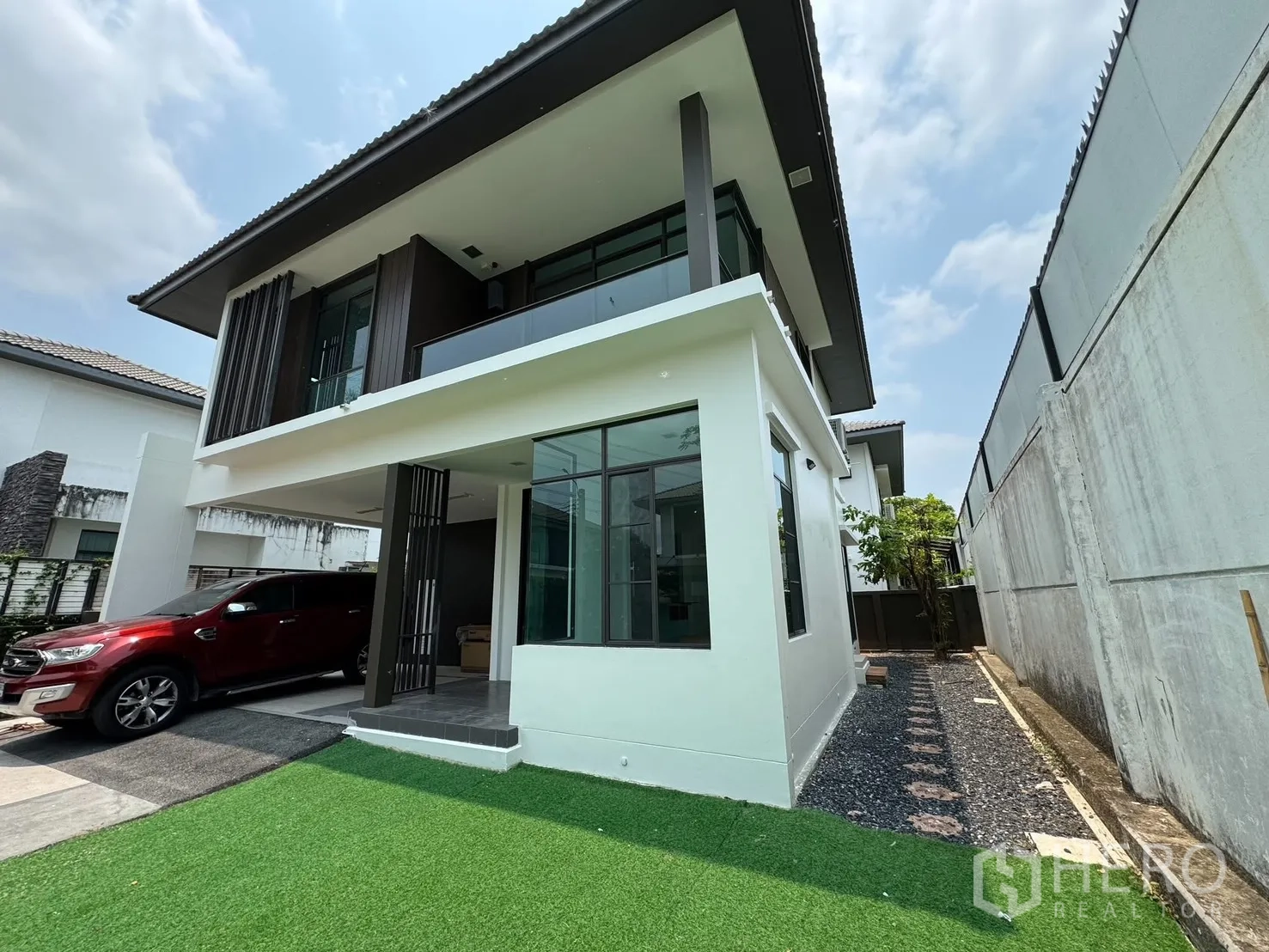 Detached House for rent in Prawet, Bangkok - Angled exterior view showing a glass balcony, large windows and a gravel path.