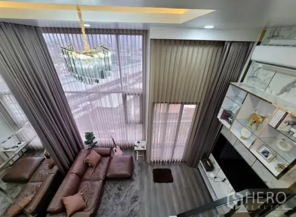 Condominium for rent in Chatuchak, Bangkok - Double-height living room with sheer floor-to-ceiling curtains, crystal chandelier, brown leather sofa, marble display shelving and city view.