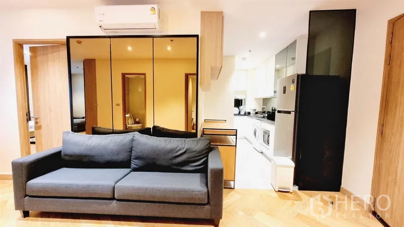 Condominium for sale in Watthana, Bangkok - Cozy living area with dark gray sofa, mirrored wall storage, air-conditioner and an open kitchen in a Watthana condominium.