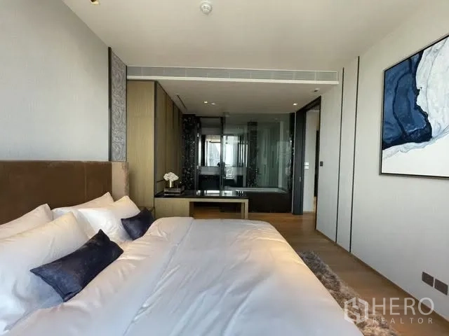 Condominium for rent or sale in Khlong Toei, Bangkok - Spacious bedroom with upholstered headboard, wall art, and a view into the bathroom through a glass wall.