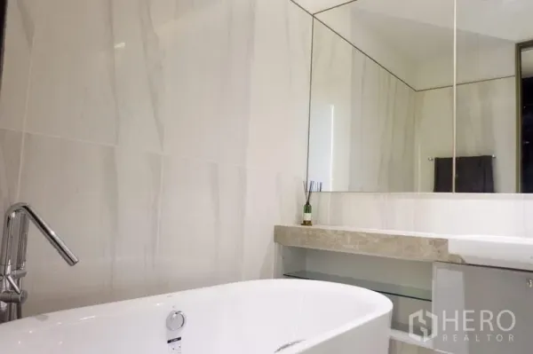 Condominium for sale in Khlong Toei, Bangkok - Modern bathroom with a freestanding bathtub, marble-style wall tiles, and a wide mirrored wall.