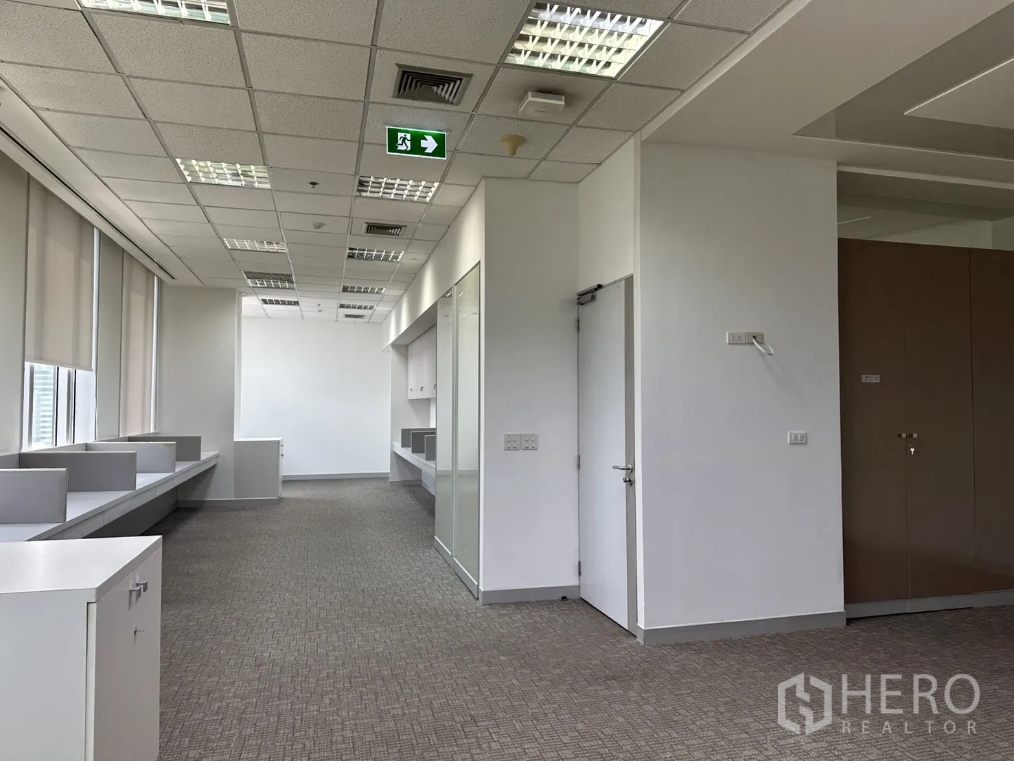 ออฟฟิศ for rent in Khlong Toei, Bangkok - โซนออฟฟิศยาวพร้อมฝ้าเพดานทีบาร์ ไฟสว่าง ป้ายทางออกฉุกเฉิน และโต๊ะทำงาน