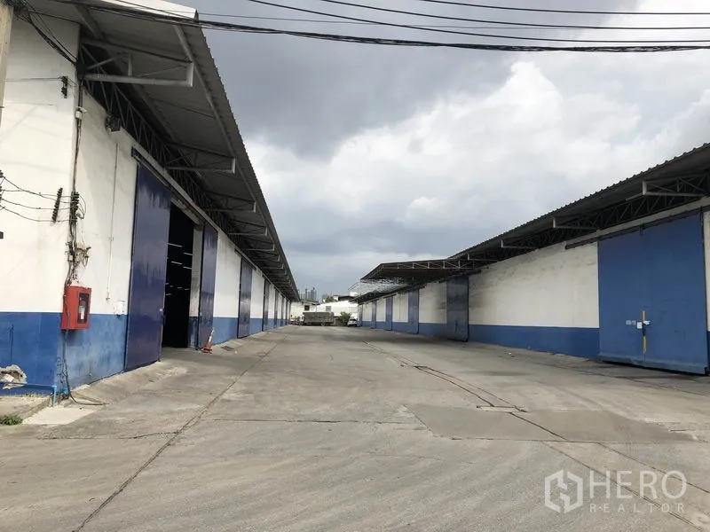 Warehouse for rent in Samut Prakan - Wide concrete driveway between blue-and-white warehouses with large sliding doors and covered eaves.