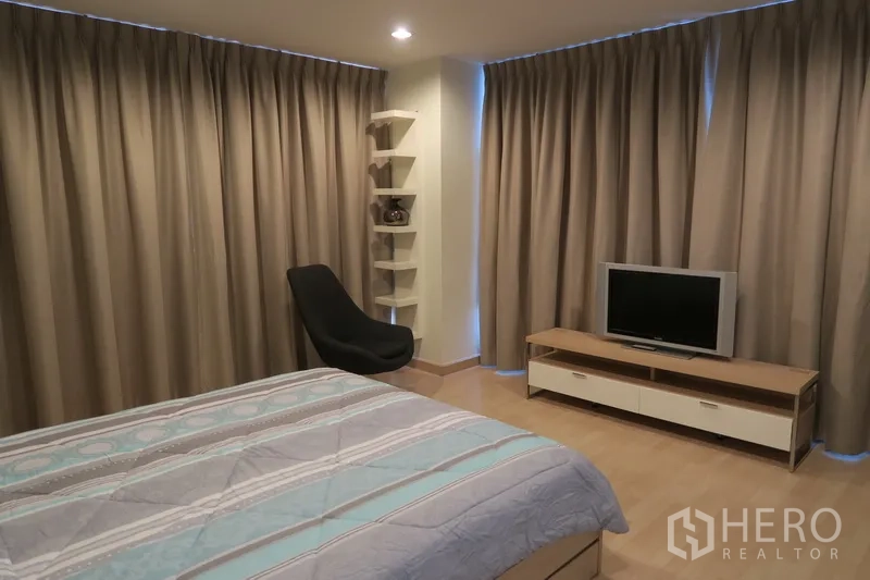Condominium for rent in Huai Khwang, Bangkok - Spacious master bedroom with TV console, corner shelves, lounge chair and full‑height curtains.