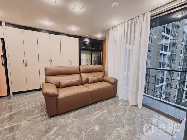 Condominium for sale in Bang Rak, Bangkok - Lounge with brown leather sofa, wall-to-wall storage and balcony glazing.