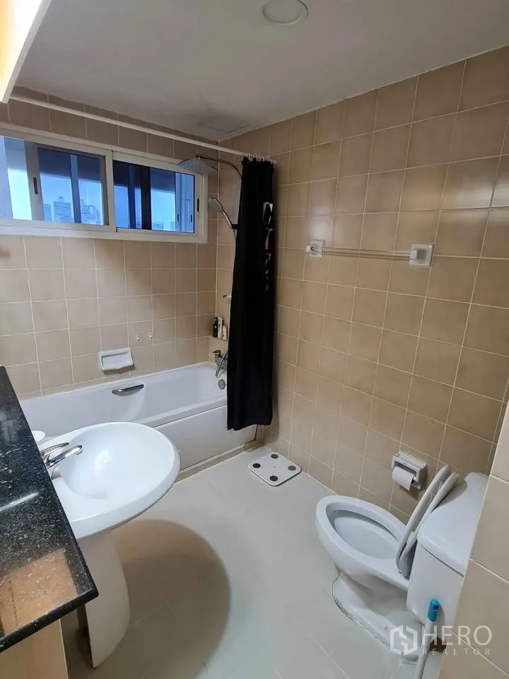 Condominium for rent in Khlong Toei, Bangkok - Tiled bathroom with bathtub, shower curtain, pedestal sink and toilet.
