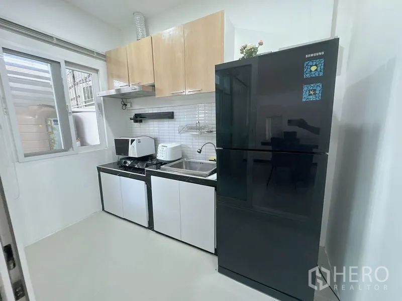 联排别墅 出租在Prawet, Bangkok - 紧凑现代厨房，黑色冰箱、灶台、水槽与吊柜临窗而设。