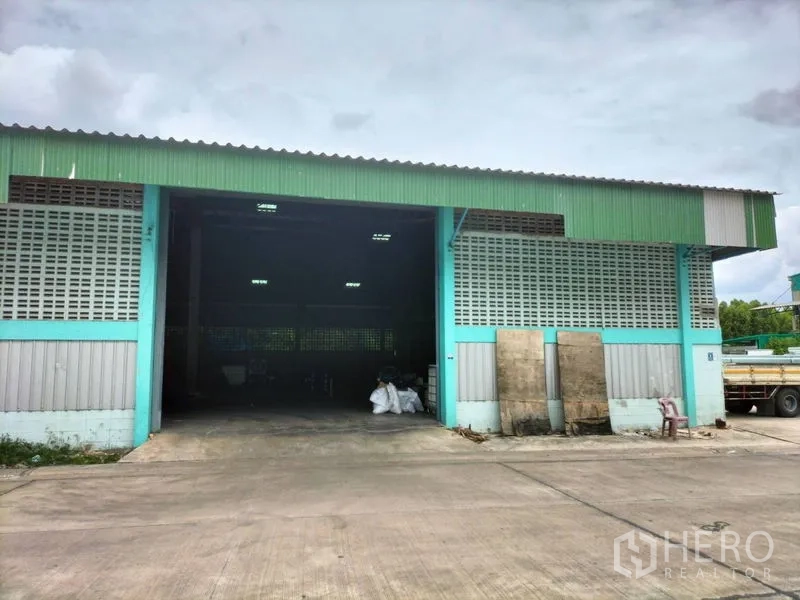 Warehouse for rent in Krathum Baen, Samut Sakhon - Front exterior of a teal and green warehouse with a wide open loading bay and concrete driveway for trucks.