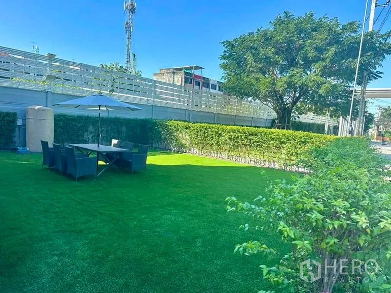 Townhouse for sale in Sai Mai, Bangkok - Large private lawn with outdoor dining table, umbrella, hedges, and a shade tree.