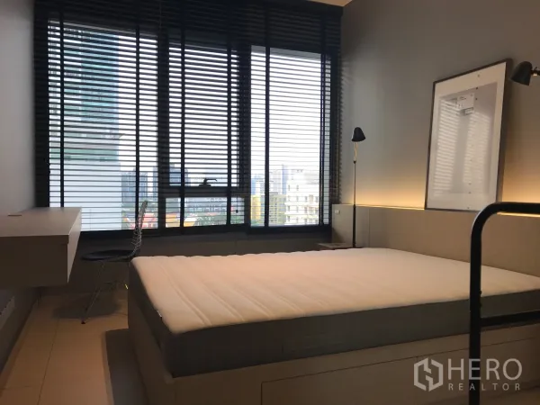 Condominium for rent in Watthana, Bangkok - Bright bedroom with large window blinds, desk and chair, and city view.
