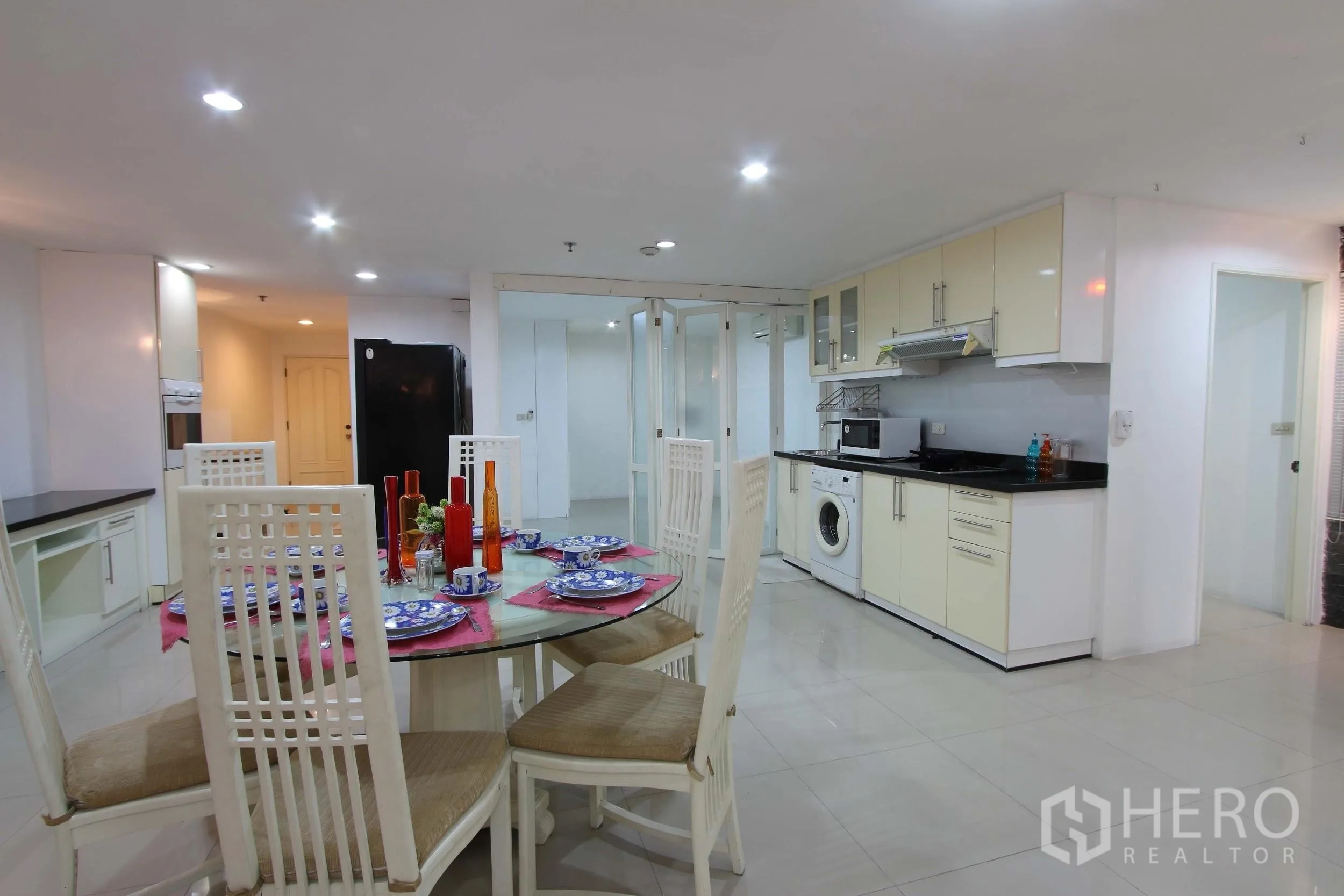 Condominium for rent in Watthana, Bangkok - Open-plan kitchen and dining area featuring white cabinets, glass-top table set for four, washing machine, and sliding glass partitions.