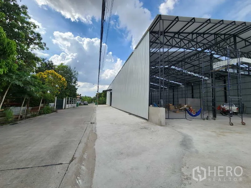 Warehouse for rent in Pak Kret, Nonthaburi - Side elevation of a long metal-clad warehouse beside a tree-lined street and wide driveway.