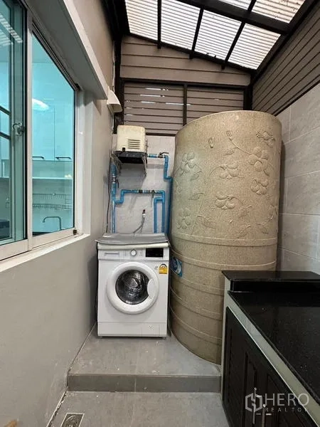 Townhouse for rent in Chom Thong, Bangkok - Laundry nook with front-loading washing machine and a large water tank under a skylight roof.