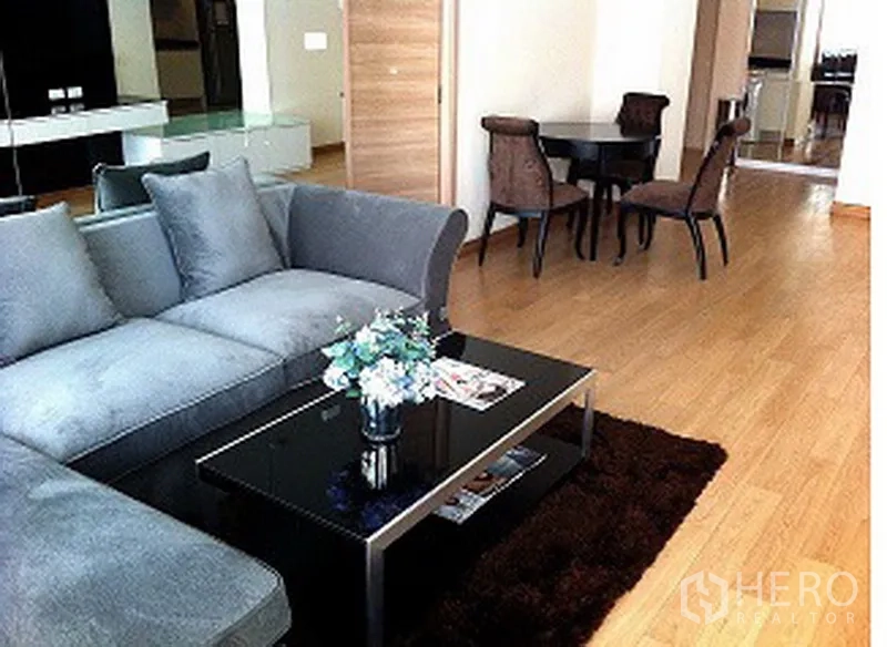 Condominium for rent or sale in Pathum Wan, Bangkok - Living room with gray sectional sofa, glass coffee table, dining set and wood floors.