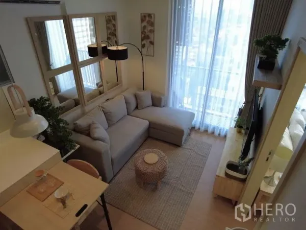 Condominium for sale in Watthana, Bangkok - Bright living room with an L-shaped sofa, mirrors, floor lamp, and balcony with sheer curtains overlooking the city.