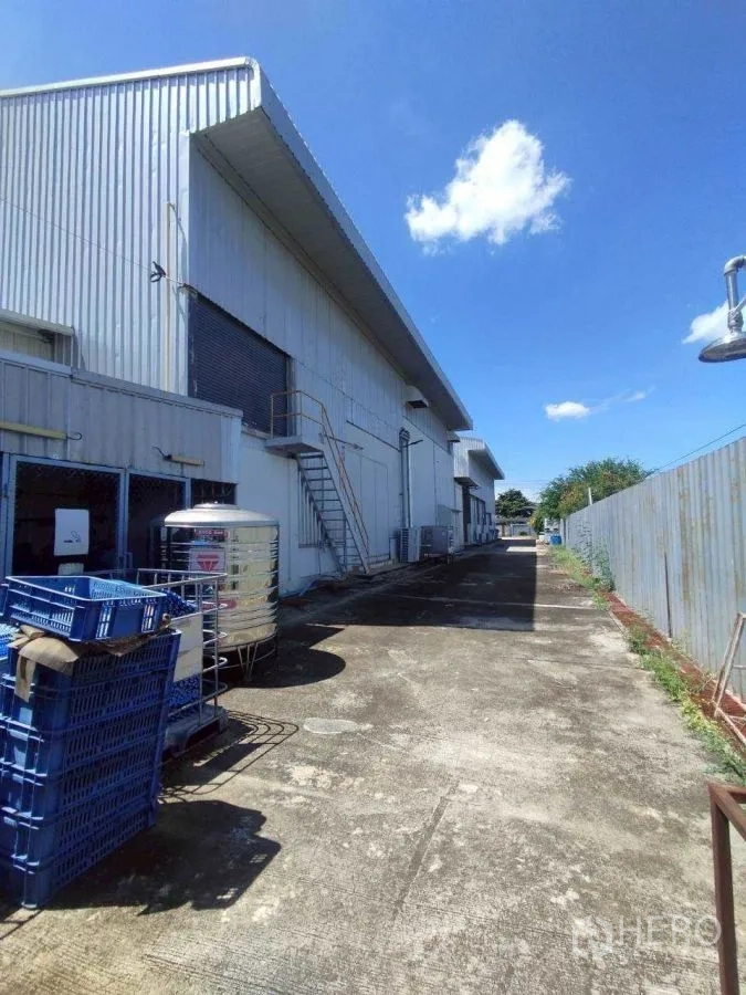 Factory for rent or sale in Mueang Nakhon Pathom, Nakhon Pathom - Side view of the metal factory with roller door, external stair, and storage crates.