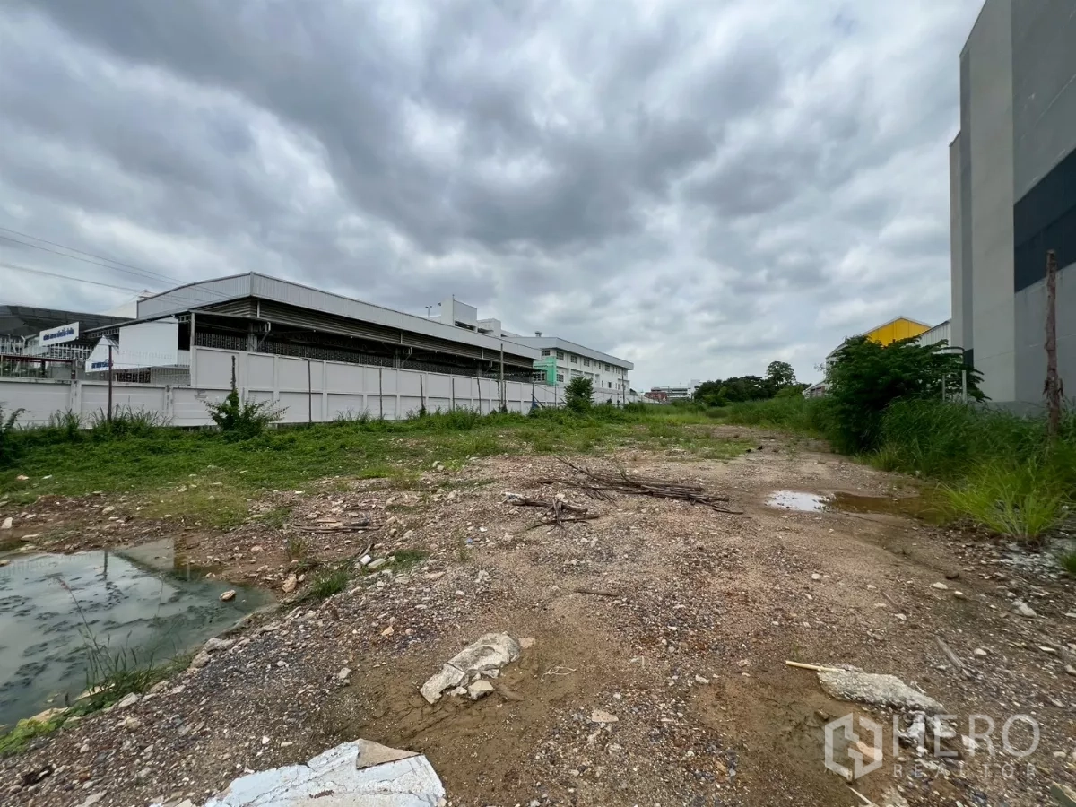 Land for sale - Open plot with rough soil and drainage beside modern warehouse complexes in a logistics area.