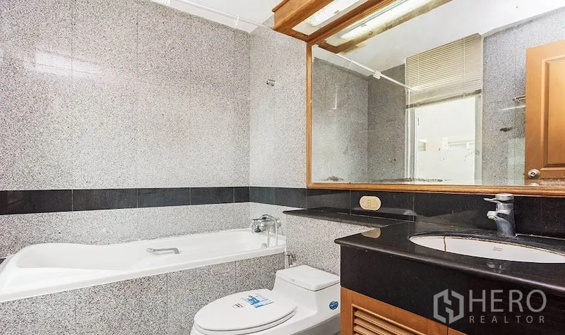 Condominium for rent in Pathum Wan, Bangkok - Modern bathroom with granite tiles, bathtub with shower, large mirror, and vanity with black countertop.