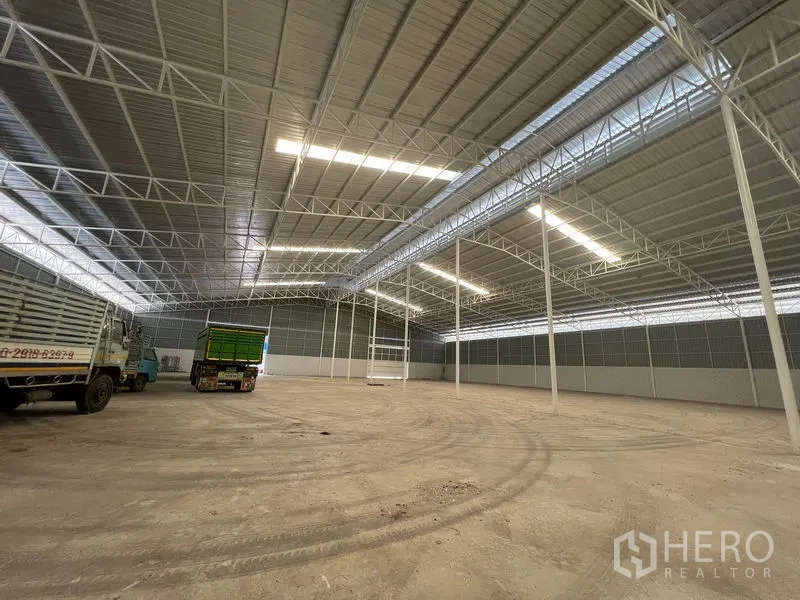 Warehouse for rent in Lat Krabang, Bangkok - Wide warehouse interior with skylights and trucks maneuvering on a concrete floor.