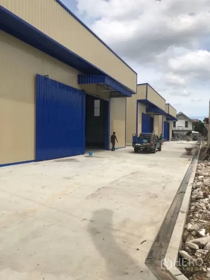 Warehouse for rent in Wang Thonglang, Bangkok - Modern row of warehouses with blue doors and a wide concrete driveway for loading and parking.