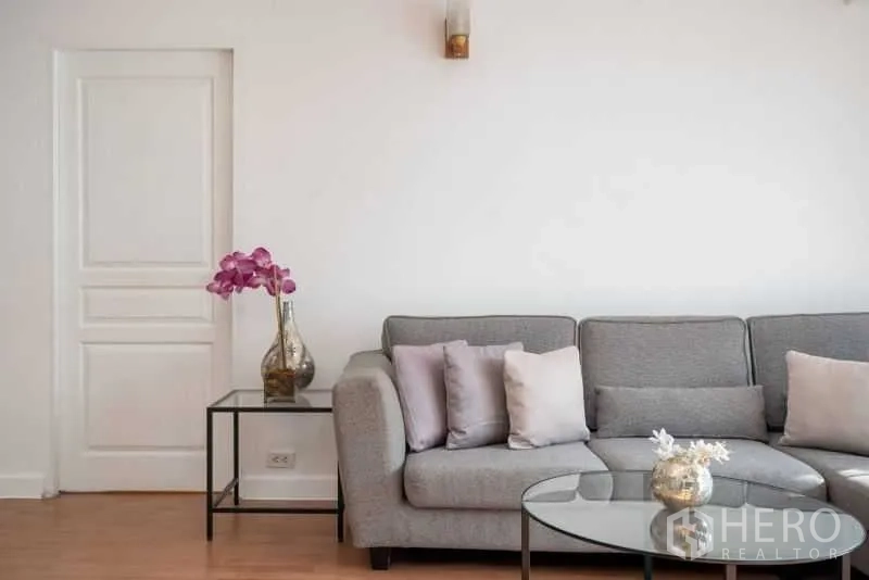 公寓 出租在Khlong Toei, Bangkok - 近景展示灰色转角沙发、玻璃茶几与兰花装饰。