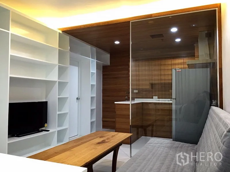 Condominium for rent in Bang Rak, Bangkok - Cozy living area facing a TV unit, with a glass wall to the wood-finished kitchen and recessed ceiling lights.