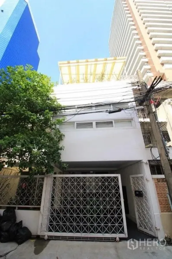 ทาวน์เฮาส์ ให้เช่าในWatthana, Bangkok - ด้านหน้าทาวน์เฮ้าส์สีขาวพร้อมประตูรั้วเหล็กลาย ข้างต้นไม้และอาคารสูงโดยรอบ