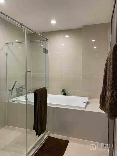 公寓 出租在Bang Na, Bangkok - 现代浴室，玻璃淋浴房与嵌入式浴缸相邻，米色瓷砖。