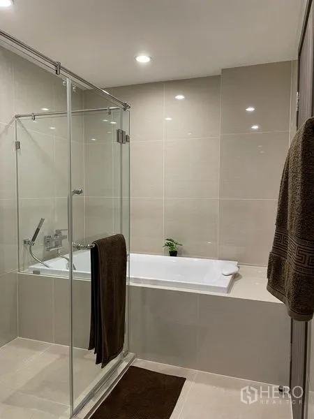 公寓 出租在Bang Na, Bangkok - 现代浴室，玻璃淋浴房与嵌入式浴缸相邻，米色瓷砖。