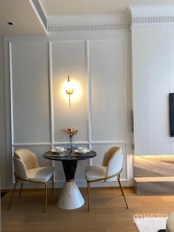 Condominium for rent in Pathum Wan, Bangkok - Compact dining nook with a round marble table, two upholstered chairs, and a wall sconce.