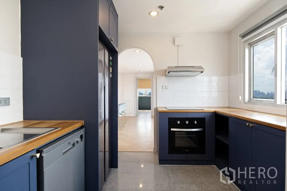 คอนโด ขายในBang Na, Bangkok - ครัวมีหน้าต่าง ตู้สีน้ำเงิน ท็อปไม้ เตาอบบิวท์อิน และซุ้มโค้งเชื่อมสู่ห้องทานอาหาร