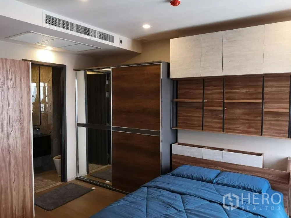 Condominium for rent in Watthana, Bangkok - Bedroom with blue bedding, built-in wooden shelves, and a mirrored sliding wardrobe beside the bathroom entrance.