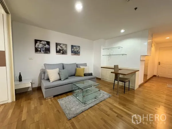 Condominium for rent in Khlong Toei, Bangkok - Bright living room with grey sofa, glass coffee table, wood floors and built‑in desk.