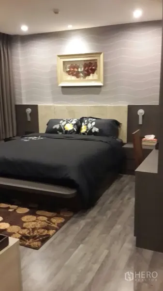 Condominium for rent in Mueang Samut Prakan, Samut Prakan - Cozy bedroom with king bed, soft ceiling lights, wood flooring, and a built-in desk with framed artwork on the feature wall.