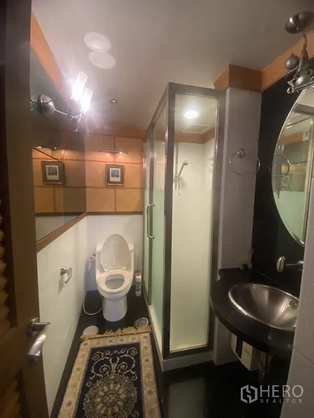Condominium for rent in Pathum Wan, Bangkok - Bathroom with glass-enclosed shower, round mirror and stainless sink beside the toilet.