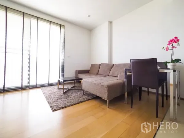 公寓 出租在Watthana, Bangkok - 客厅设L形沙发、地毯、茶几与餐桌，旁有高窗。