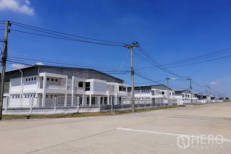 Warehouse for rent in Phra Nakhon Si Ayutthaya - Row of modern white warehouse buildings with perimeter fence along a wide industrial road under a clear blue sky in Uthai, Ayutthaya.