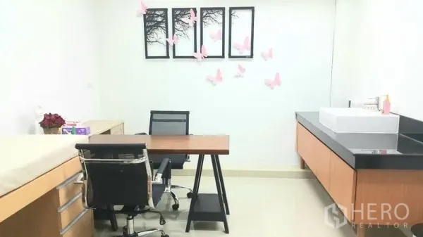 Shophouse for rent in Khan Na Yao, Bangkok - Bright consultation room with wooden desk, black office chair, examination bed, wall art and a countertop sink.