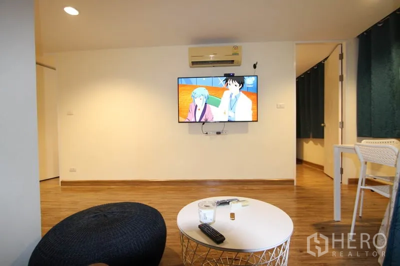 Condominium for rent or sale in Wang Thonglang, Bangkok - Living room with wall-mounted TV, ottoman, and round coffee table on wood-look floors.