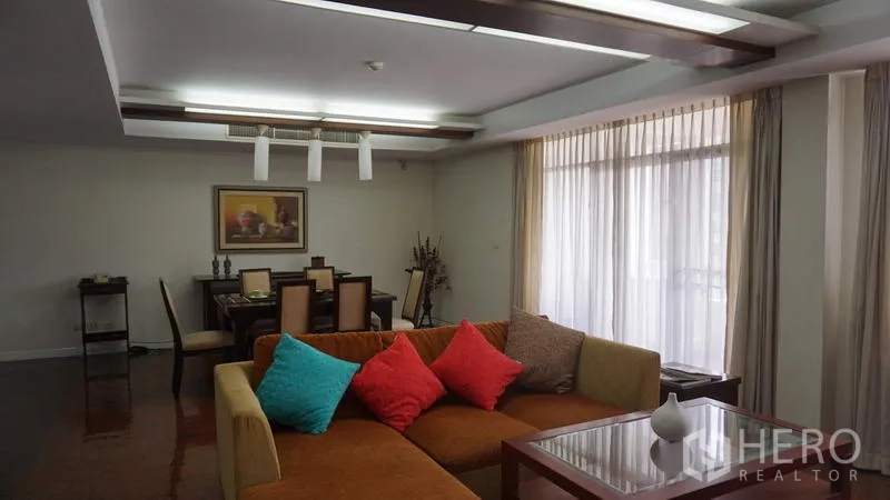 房间 出租在Khlong Toei, Bangkok - 起居区一隅，L 形沙发配彩色抱枕与茶几，旁为大面积采光的餐区。