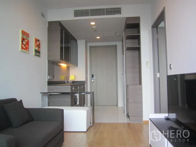 คอนโด ให้เช่าในKhlong Toei, Bangkok - ห้องนั่งเล่นสว่างพร้อมโซฟาสีเทา ชั้นวางทีวี และครัวขนาดกะทัดรัดตู้ลายมันวาว พื้นไม้