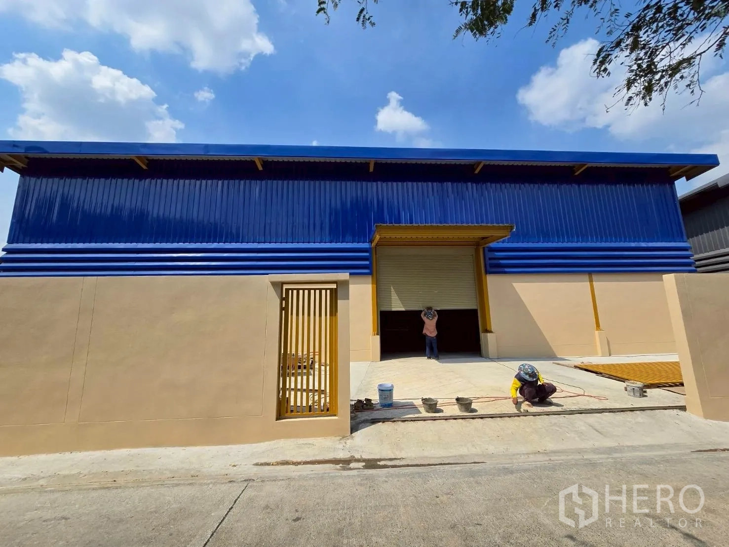 货栈 for rent in Khlong Sam Wa, Bangkok - 现代仓库正立面，蓝色板材、米色下部墙体，大卷闸门、人员门与装卸前场。