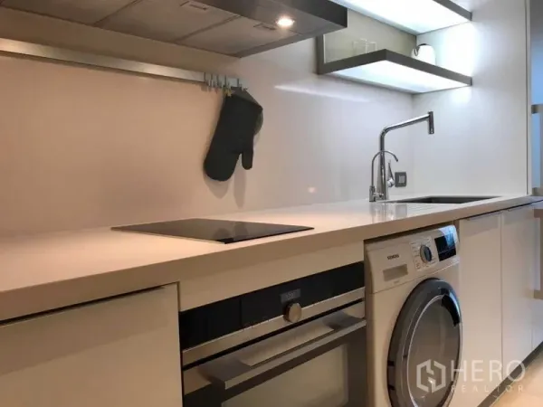 Condominium for rent in Pathum Wan, Bangkok - Modern compact kitchen with induction cooktop, oven, sink, open shelving and a front‑load washer‑dryer