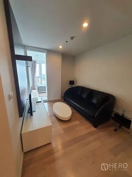 Condominium for rent in Watthana, Bangkok - Cozy living room with black sofa, TV console and wood flooring next to bedroom.