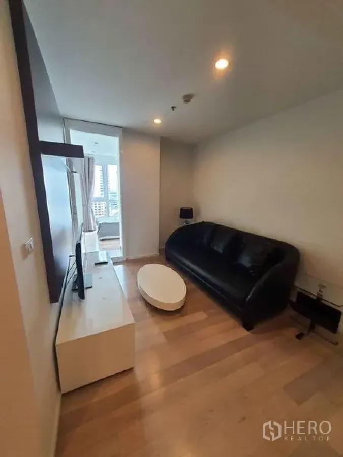 Condominium for rent in Watthana, Bangkok - Cozy living room with black sofa, TV console and wood flooring next to bedroom.