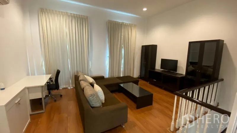 Townhouse for rent in Wang Thonglang, Bangkok - Upstairs living room with sectional sofa, TV stand, desk, and tall curtains.