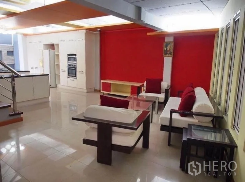 Townhouse for rent in Wang Thonglang, Bangkok - Lounge with white sofas and red cushions beside an open kitchen and a bold red feature wall.