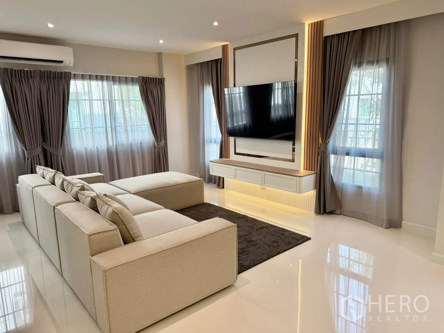 Detached House for rent in Bang Phli, Samut Prakan - Living room featuring wall-mounted TV, L-shaped sofa, glossy white floor and sheer curtains.