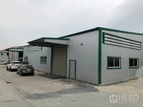 Warehouse for rent in Pak Kret, Nonthaburi - Row of metal warehouses with cars parked along a wide driveway and covered loading bay.