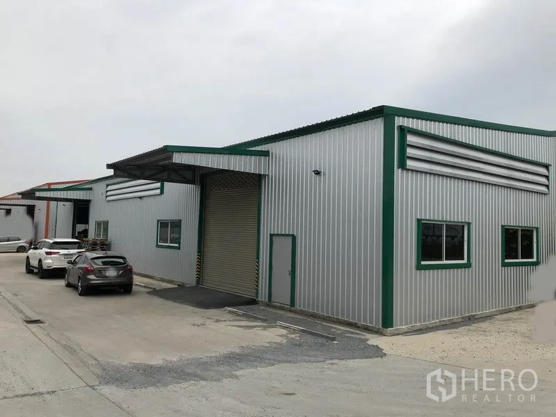 Warehouse for rent in Pak Kret, Nonthaburi - Row of metal warehouses with cars parked along a wide driveway and covered loading bay.